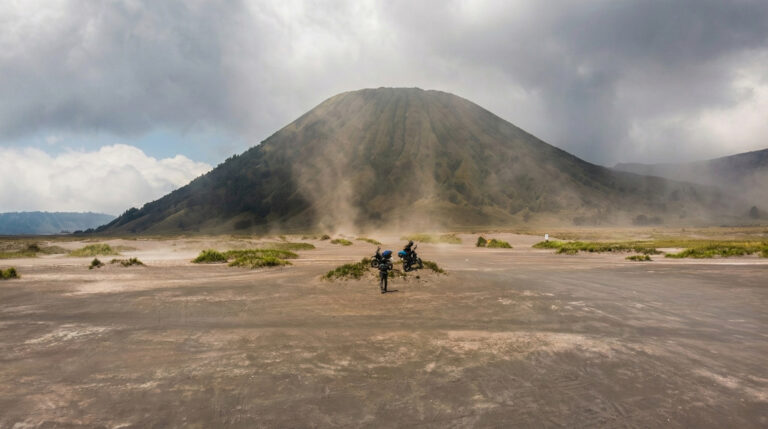 Two motorbike riders cross a dusty volcanic sand plain below a cone-shaped peak