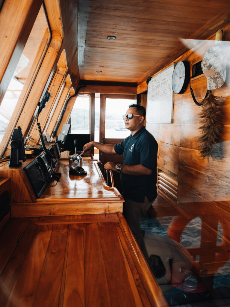 Crew member steering a phinisi from the teak wheelhouse, hand on the controls