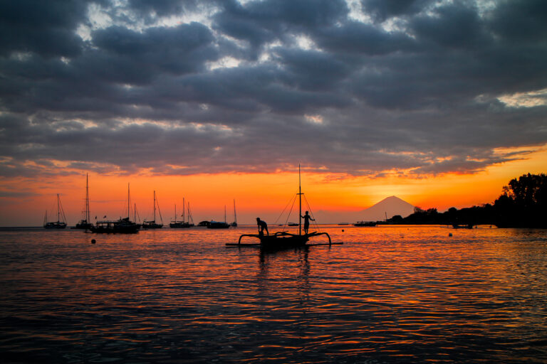 Outrigger canoe and anchored sailing yachts at sunset with a volcano on the horizon