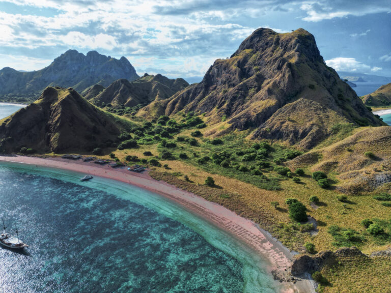 Pink-sand beach curving below Padar Island's hills with a phinisi anchored offshore, aerial view