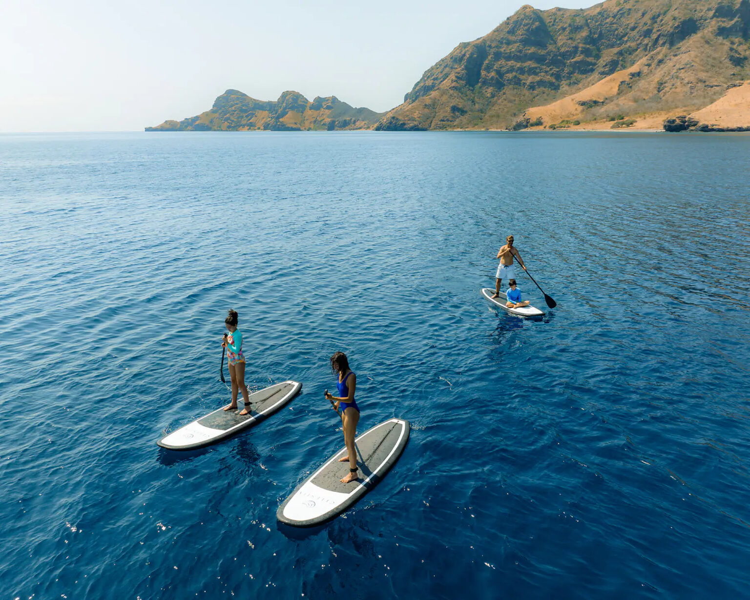 Three paddleboarders on a calm bay below dry hills, Komodo National Park, from above