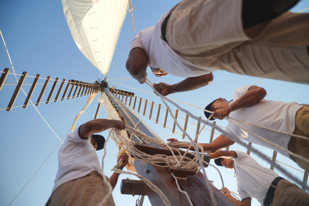 Crew raising the sail on Papiton Allo seen from below against a blue sky