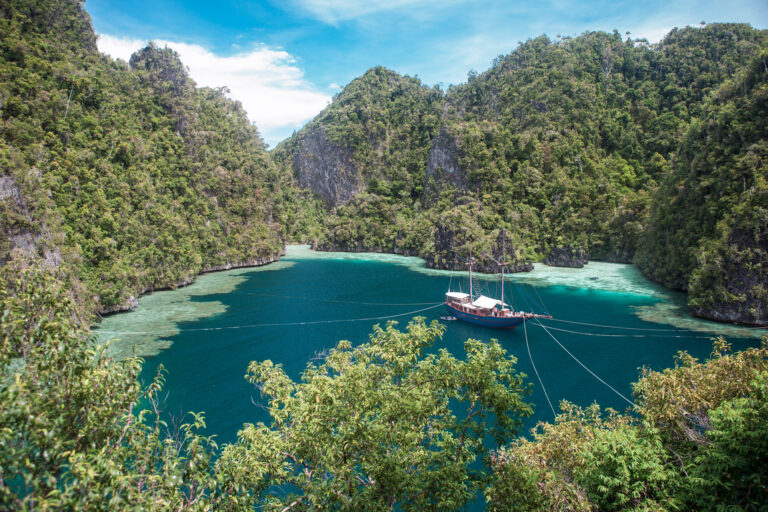 Phinisi anchored with shore lines in a sheltered lagoon of forested karst walls, Raja Ampat
