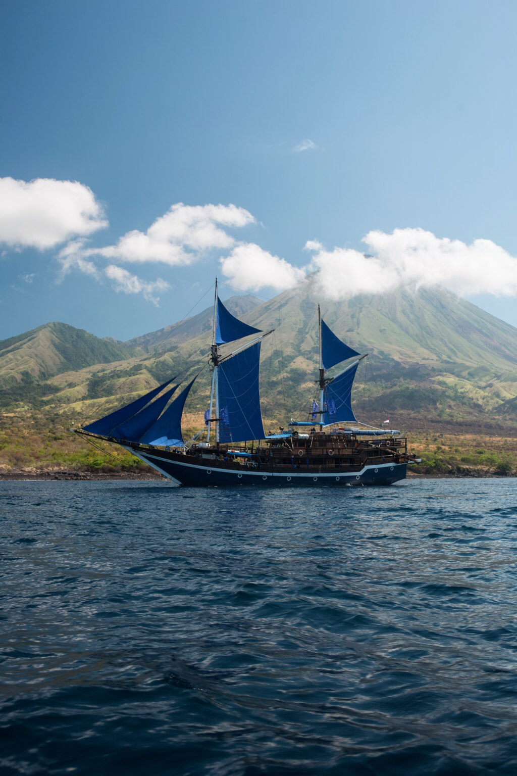 Phinisi with blue sails set below a cloud-topped volcano in Komodo waters