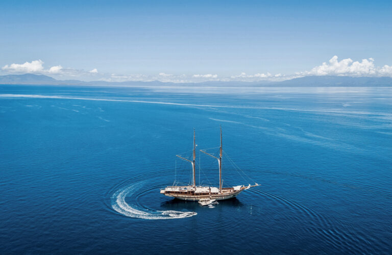 Phinisi on open blue water with a tender leaving a wake, seen from above
