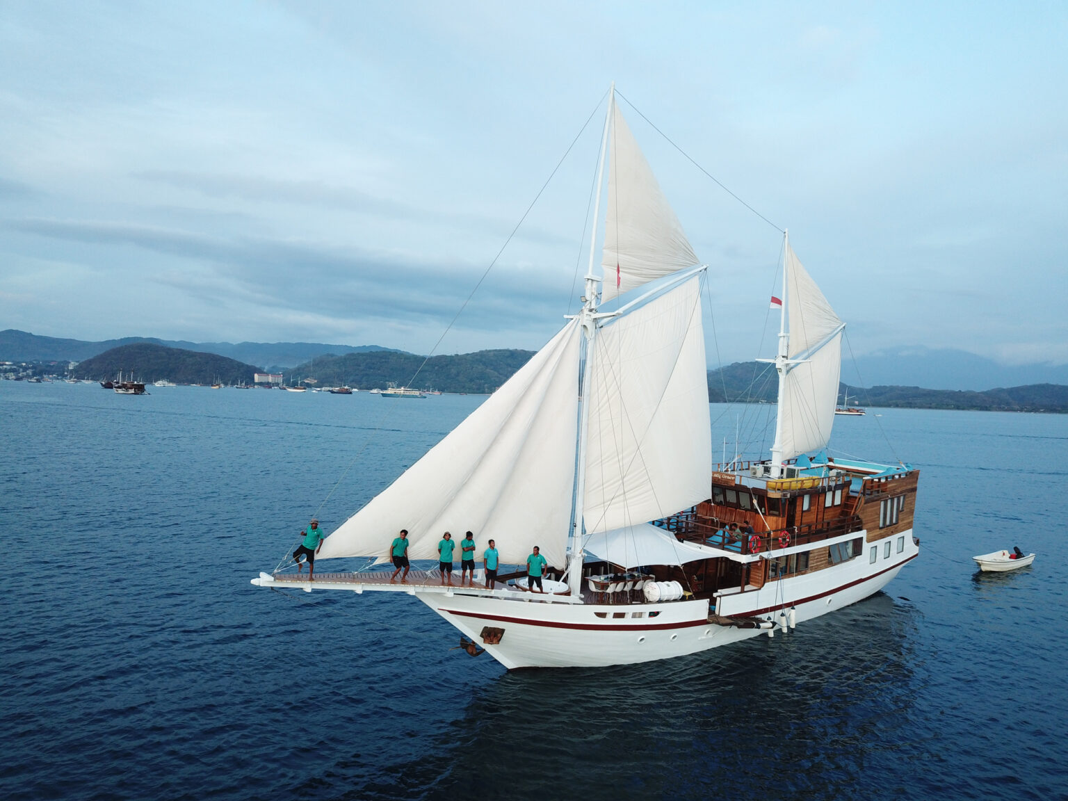 Phinisi under white sails with travelers on deck crossing a calm bay