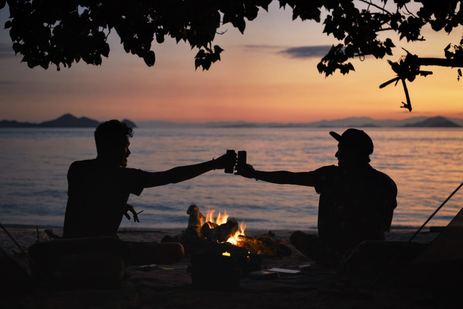 Two silhouetted friends clinking cans over a beach campfire at sunset, islands on the horizon