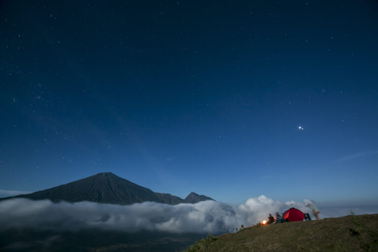 Red tent on a ridge above the clouds under a starry sky, volcano beyond