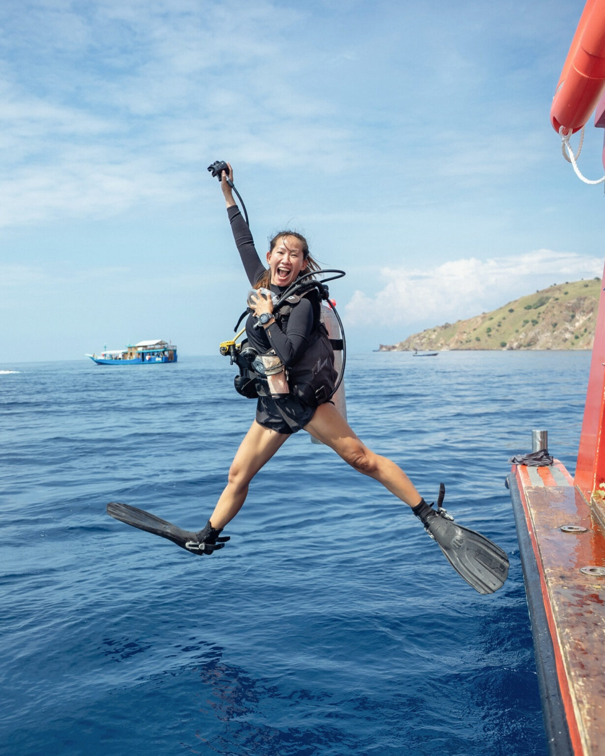 Laughing scuba diver mid giant-stride off a liveaboard, dry hills in the background, Komodo