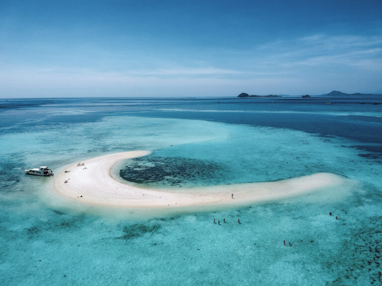 Taka Makassar sandbar curving through turquoise shallows with a speedboat moored, aerial view